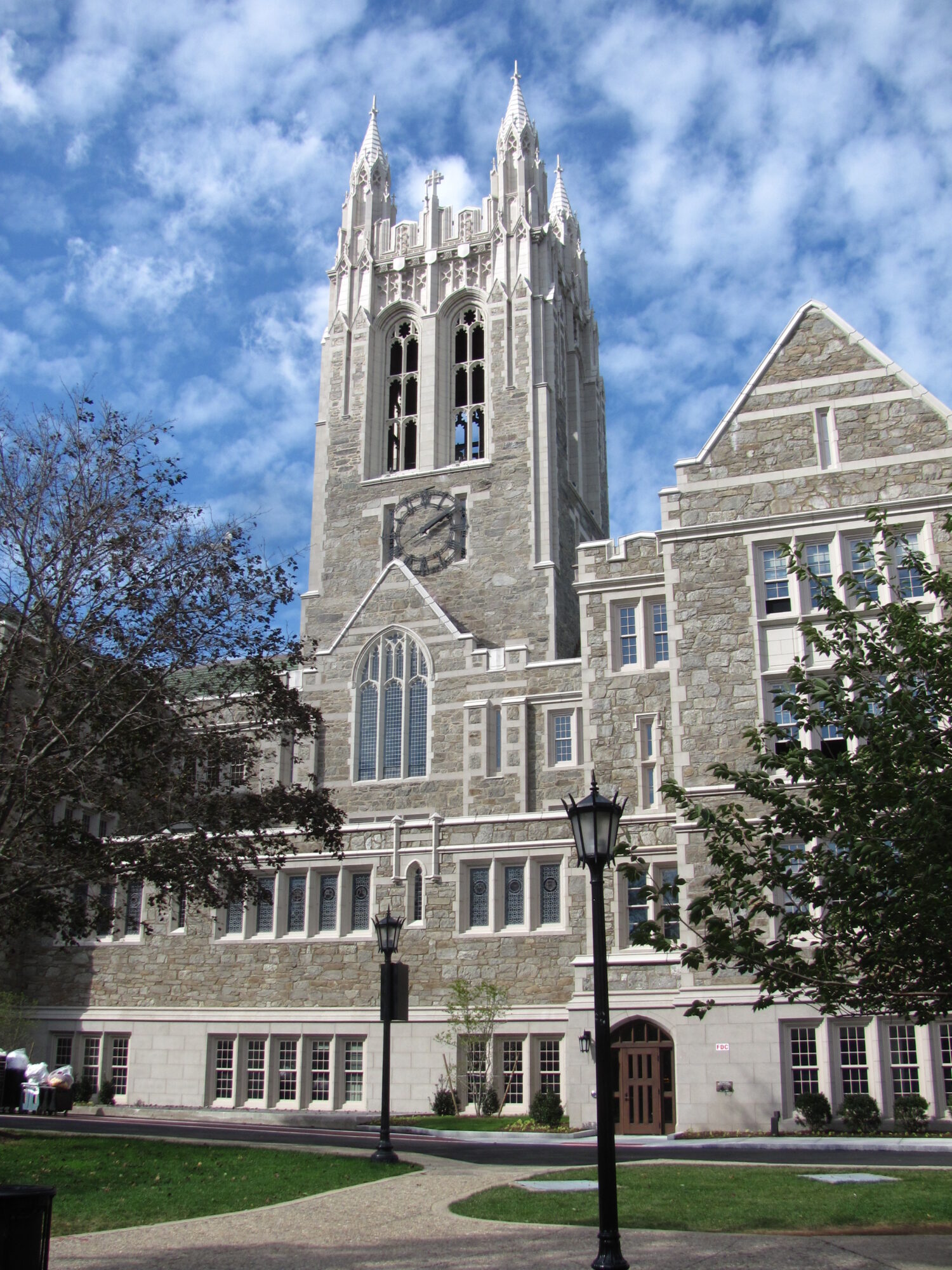 The stone Gothic tower of Gasson Hall at Boston College under a blue sky with a lamp post in the foreground