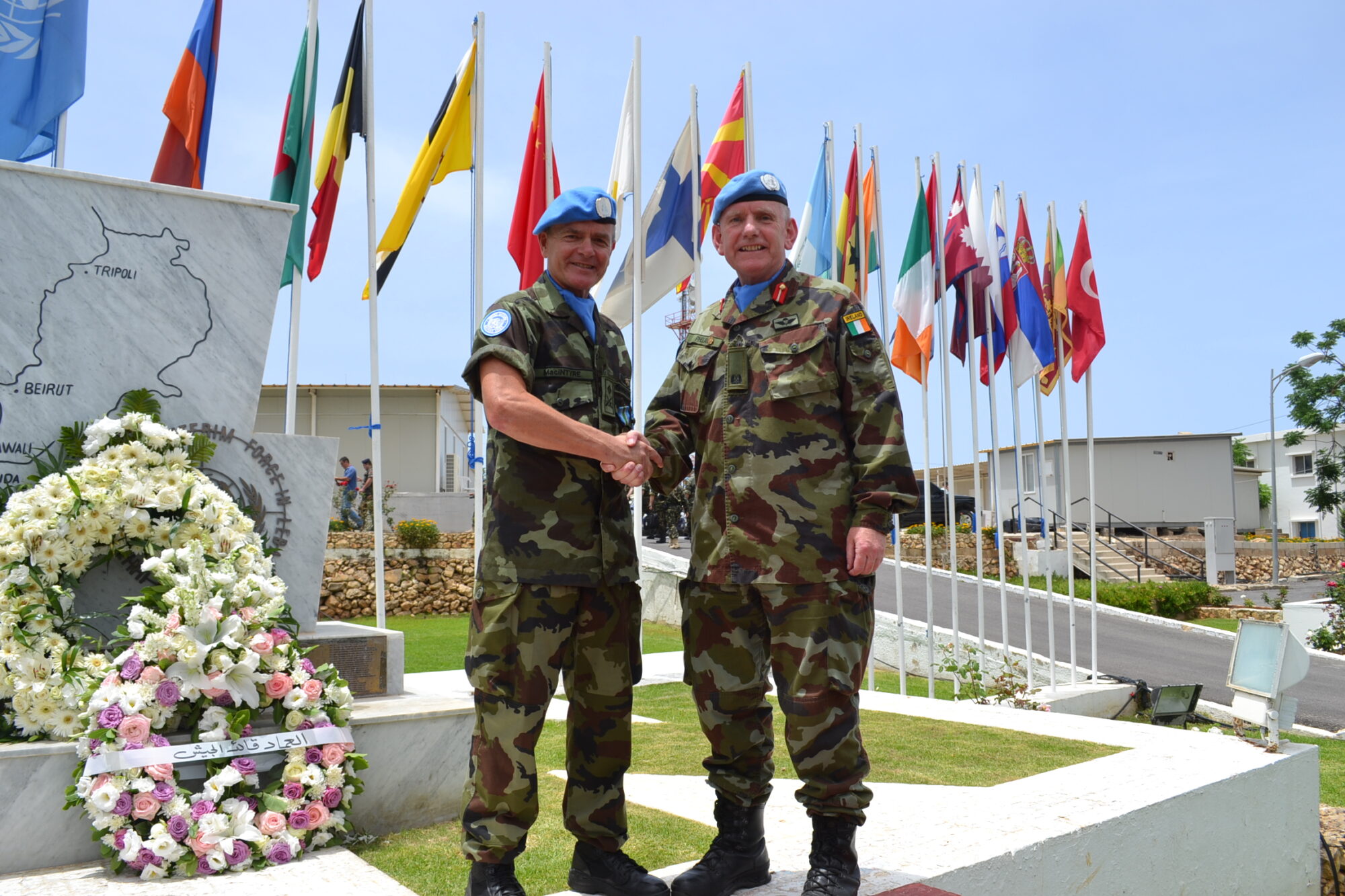 Two officers in blue UN berets shake hands in front of a floral wreath and a row of national flags at a UN peacekeeping memorial in Lebanon