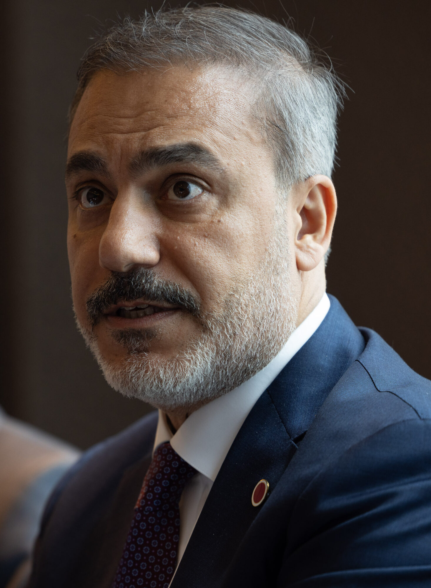 Close portrait of Hakan Fidan, a grey-haired man with a trimmed beard in a navy suit, speaking