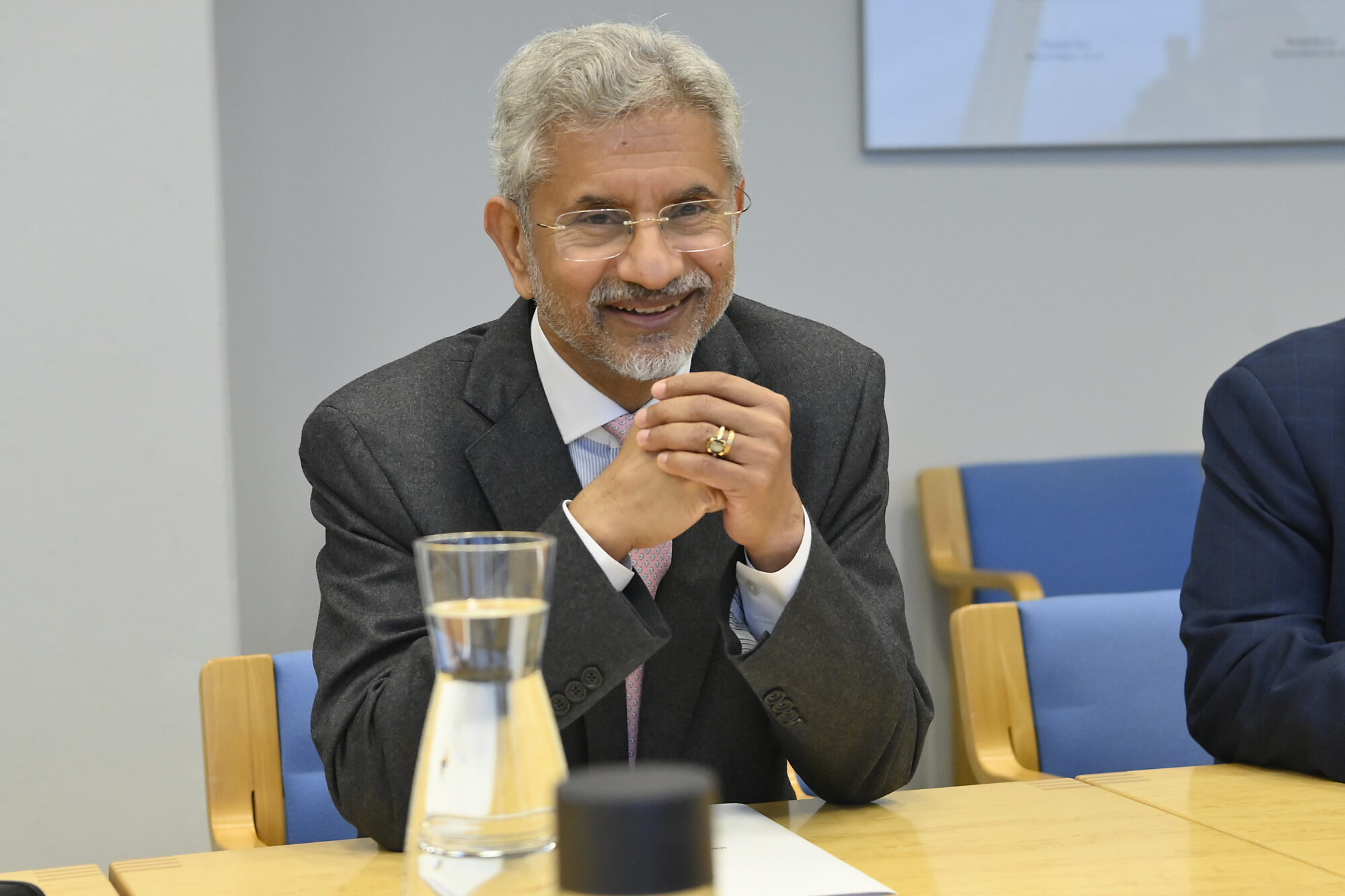 Smiling grey-haired man in rimless glasses and a grey jacket with hands clasped at a meeting table