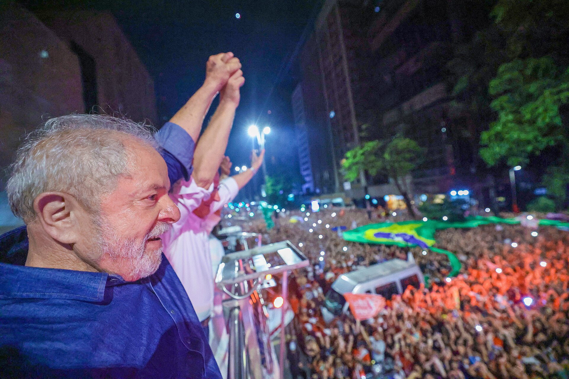 Lula, a grey-haired bearded man in a blue shirt, raises clasped hands above a large night-time crowd with a Brazilian flag