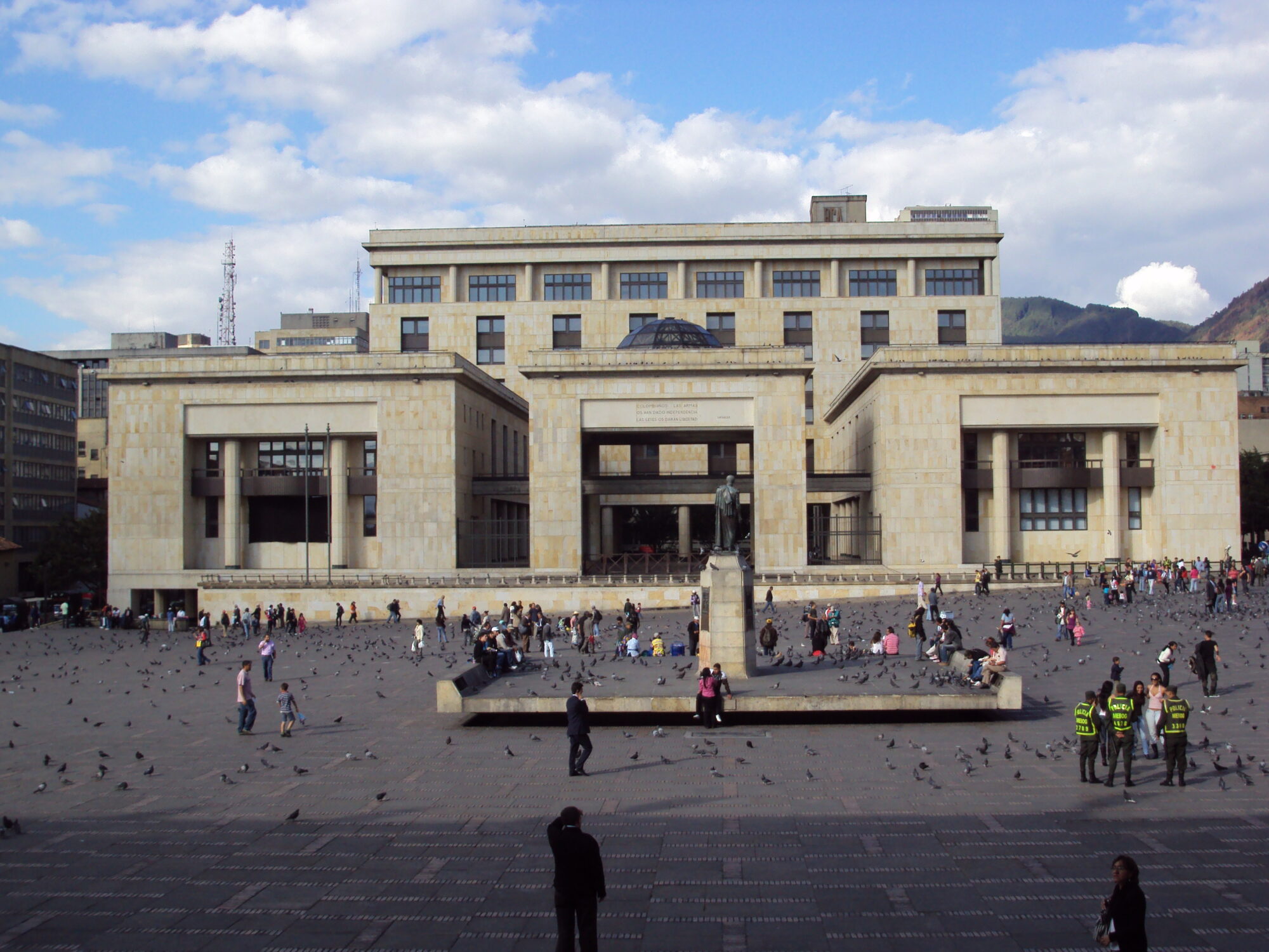 A wide paved plaza with pigeons and visitors in front of a large stone government building in Bogotá with mountains behind