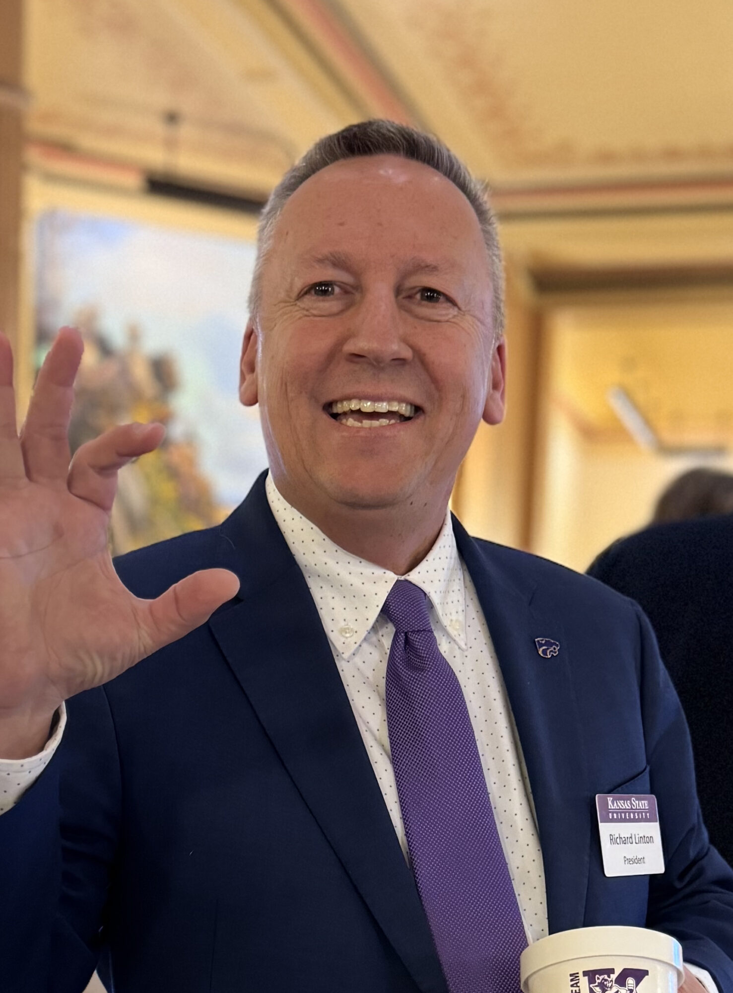Smiling man in a navy suit and purple tie with a Kansas State University name badge reading Richard Linton, President, raising one hand in greeting